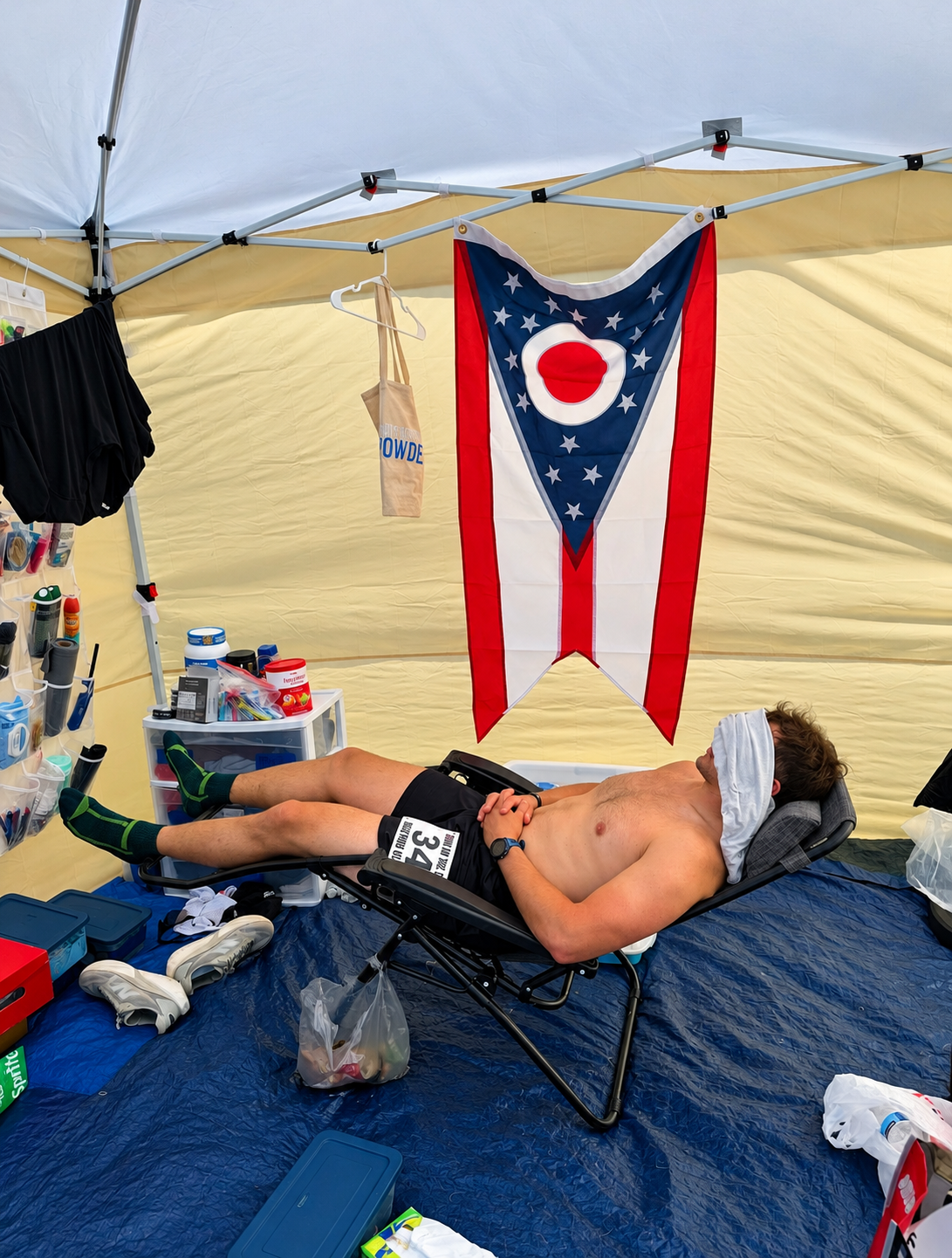 Adam Hettel resting in a reclined chair in the crew tent between loops at a backyard ultra, a towel over his face and an Ohio flag hanging overhead