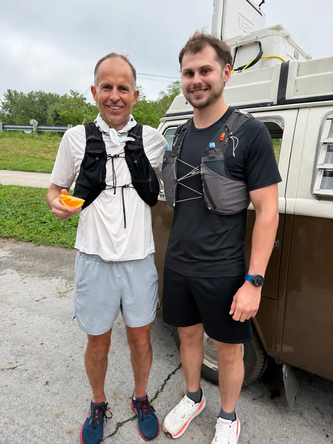 Two runners wearing hydration vests standing beside a camper van